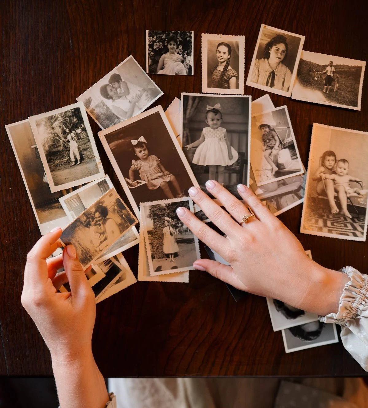 A top-down view of a person's hands sorting through scattered vintage sepia-toned photographs on a dark wooden table.
