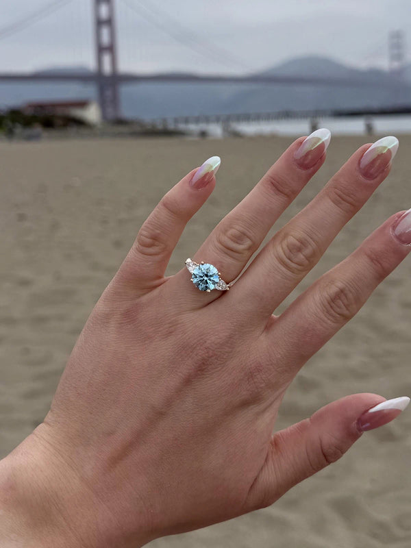 Hand wearing a blue gemstone ring with a blurred beach and bridge in the background