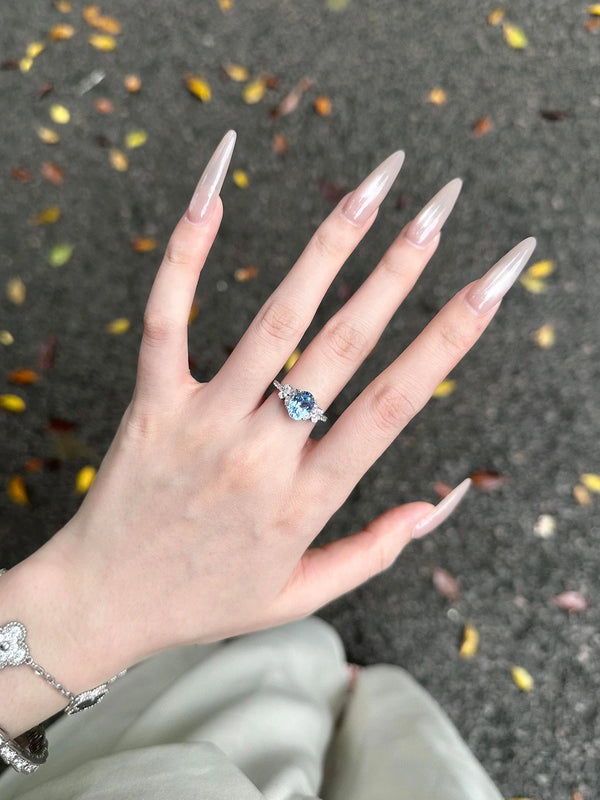 Hand with long nails wearing a ring with a blue gemstone on a dark surface with scattered leaves.