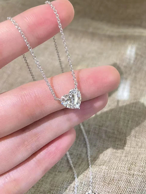 Heart-shaped diamond pendant on a chain held by a hand against a textured beige background