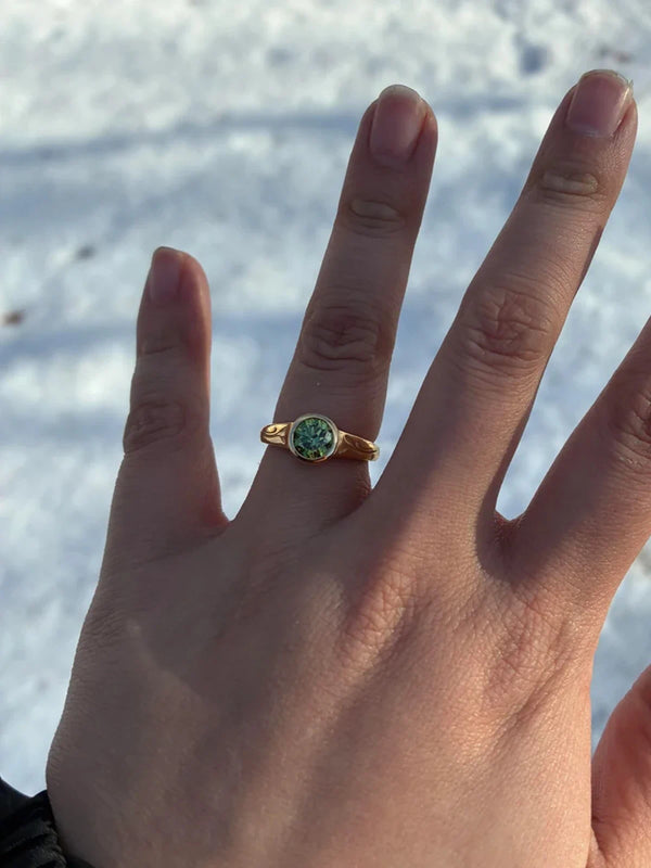 Hand wearing a gold ring with a green gemstone against a snowy background