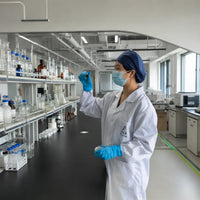 Person in a laboratory wearing a white coat, blue gloves, and a face mask, holding a test tube.